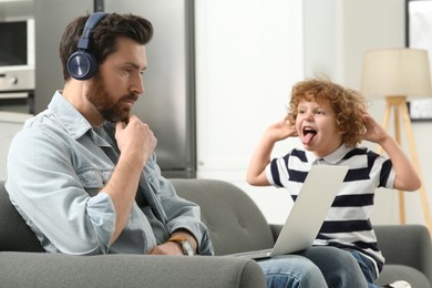 Photo of Little boy bothering his father at home. Man with laptop and headphones working remotely
