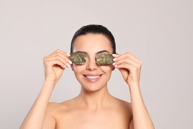 Beautiful young woman putting green tea bags on eyes against light grey background