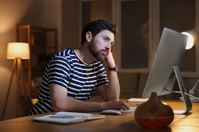 Photo of Home workplace. Tired man working with computer at wooden desk in room at night