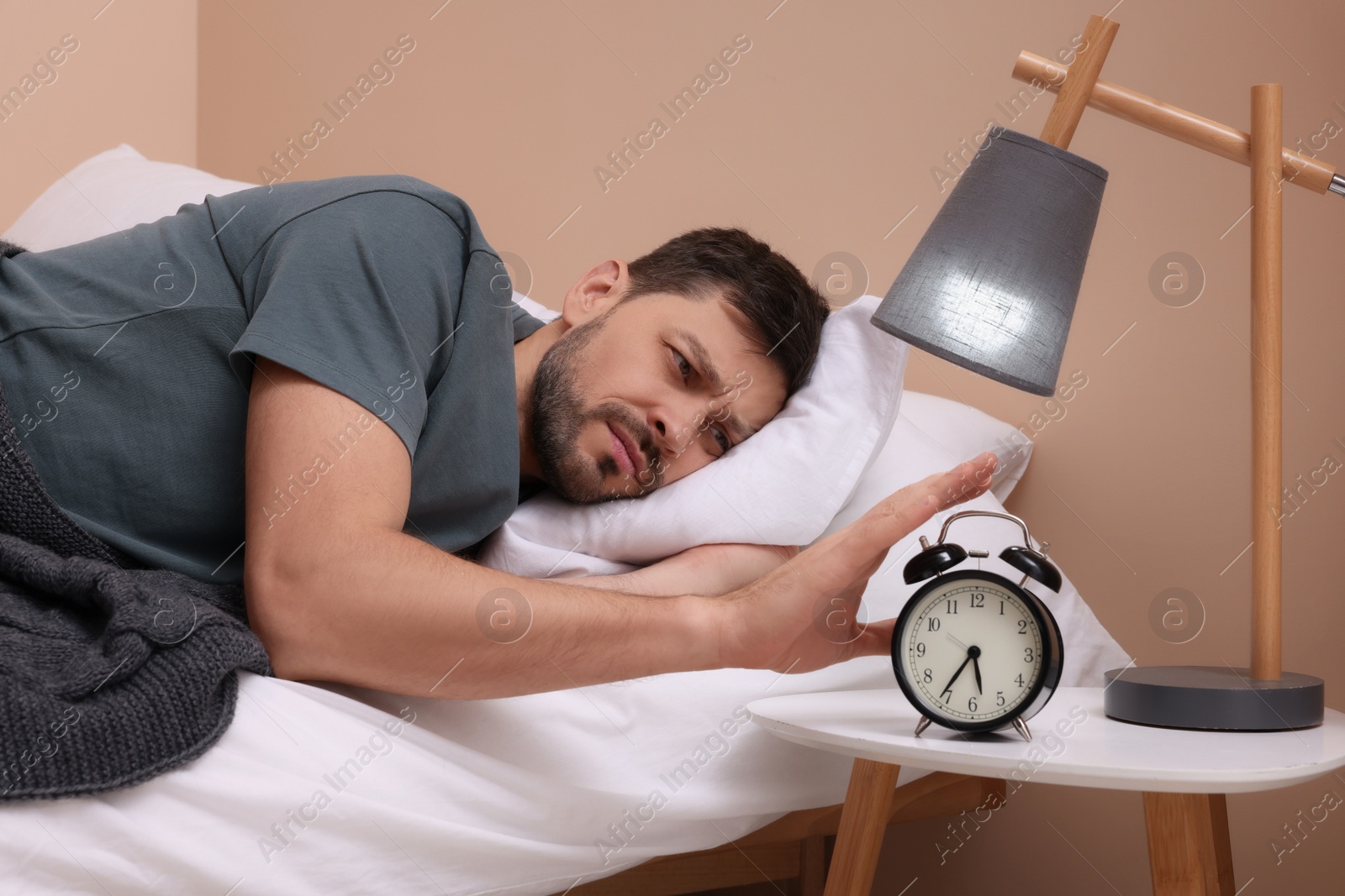 Photo of Sleepy man turning off alarm clock on nightstand in morning