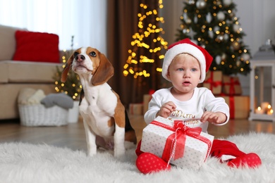 Baby in Santa hat and cute Beagle dog with gift at home against blurred Christmas lights
