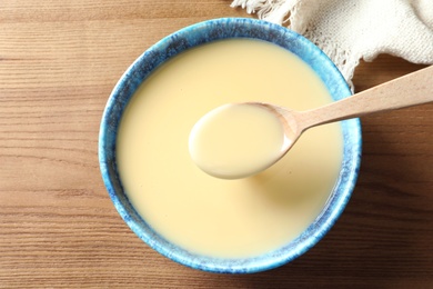 Photo of Spoon and bowl of condensed milk on wooden table, top view. Dairy products