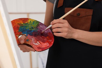 Photo of Woman mixing paints on palette with brush near easel in studio, closeup