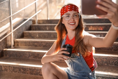 Photo of Beautiful young woman with coffee sitting on stairs and taking selfie outdoors, space for text