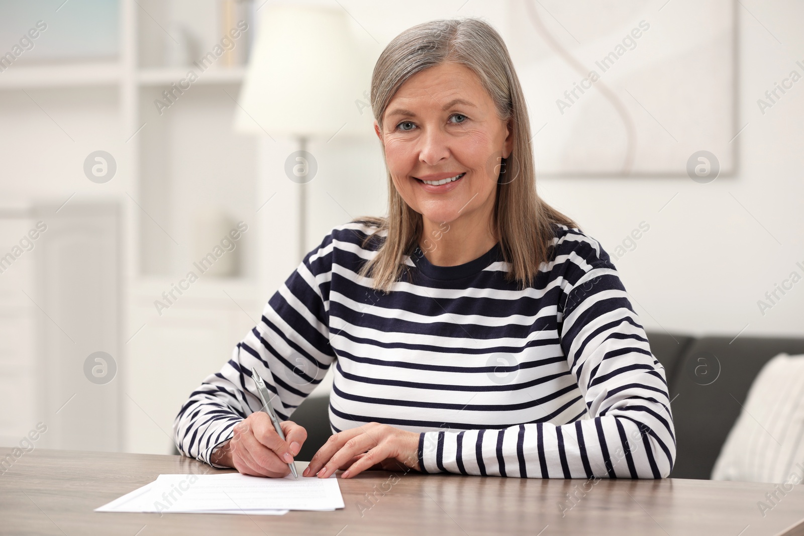 Photo of Smiling senior woman signing Last Will and Testament at wooden table indoors