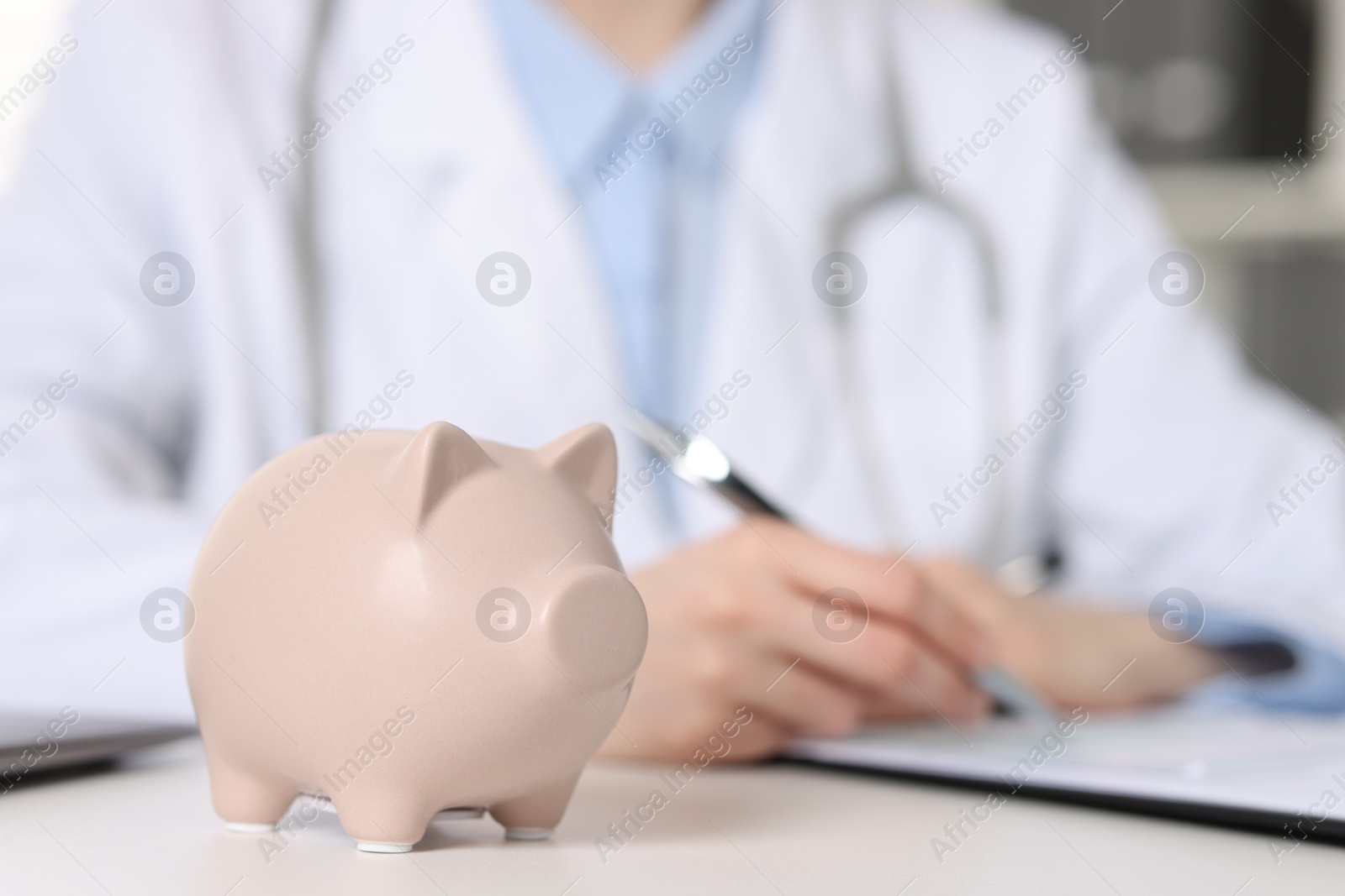 Photo of Doctor making notes at white table indoors, focus on piggy bank