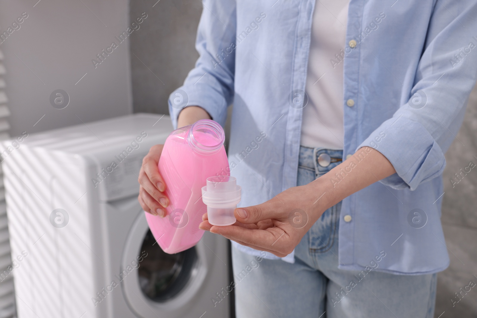 Photo of Woman pouring fabric softener from bottle into cap for washing clothes indoors, closeup