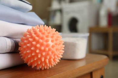 Photo of Orange dryer ball and detergent near stacked clean clothes on wooden table in laundry room, closeup. Space for text