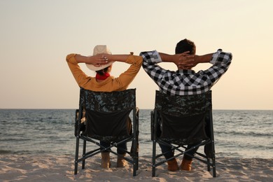Photo of Couple sitting in camping chairs and enjoying seascape on beach, back view