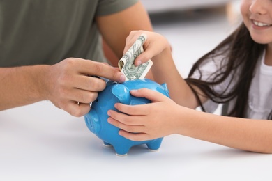 Little girl with her father putting money into piggy bank at table, closeup