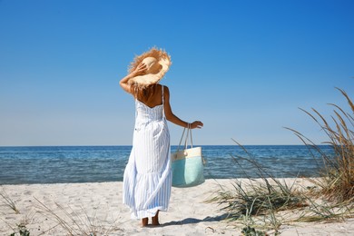 Photo of Woman with beach bag and straw hat on sand near sea, back view