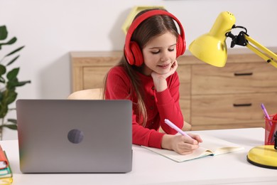 Photo of E-learning. Cute girl taking notes during online lesson at table indoors