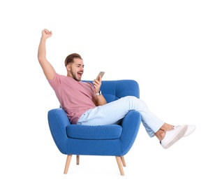 Emotional young man with smartphone in armchair on white background