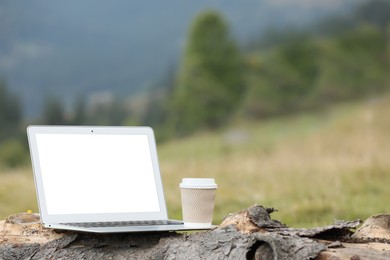 Photo of Modern laptop with blank screen and coffee cup on tree trunk in nature, space for text. Working outdoors