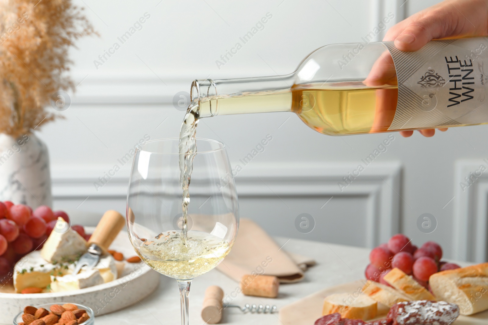 Photo of Woman pouring white wine from bottle into glass at table with snacks, closeup