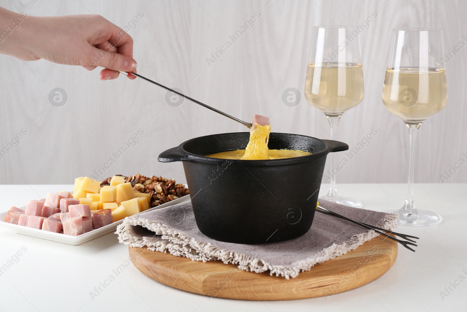 Photo of Dipping piece of ham into fondue pot with tasty melted cheese at white table, closeup