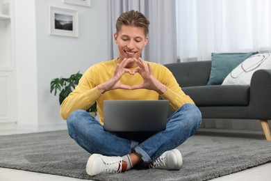 Photo of Happy young man having video chat via laptop and making heart with hands on carpet indoors. Long-distance relationship