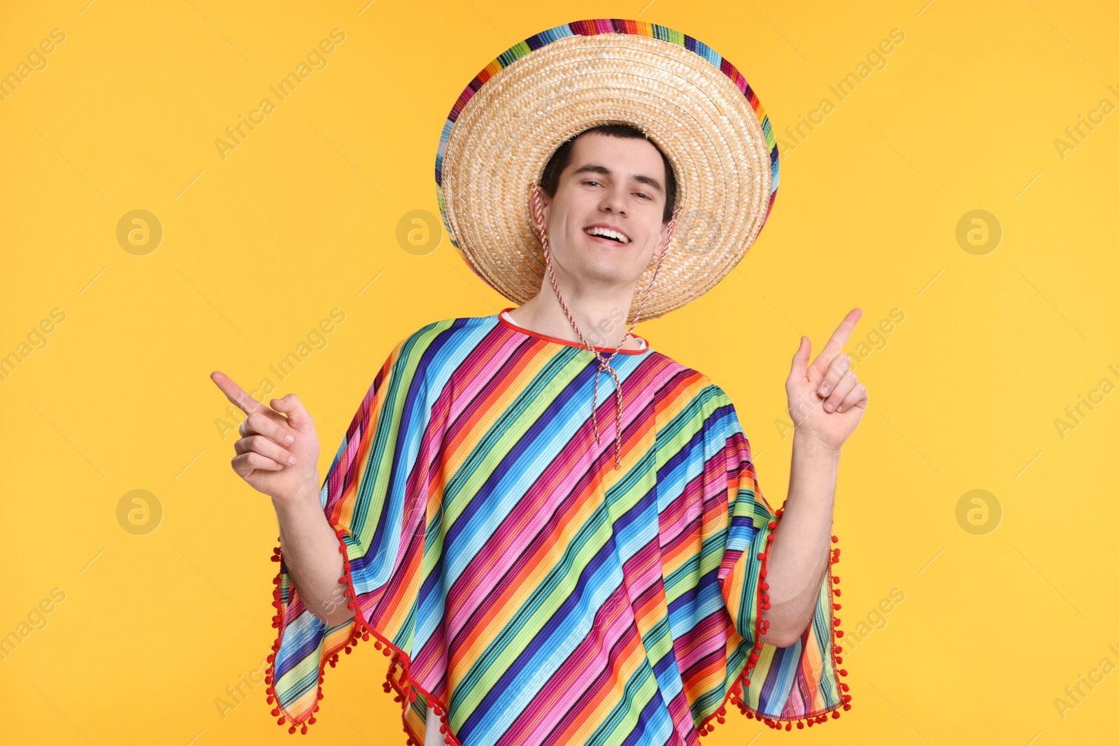 Photo of Young man in Mexican sombrero hat and poncho pointing at something on yellow background