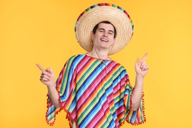 Photo of Young man in Mexican sombrero hat and poncho pointing at something on yellow background