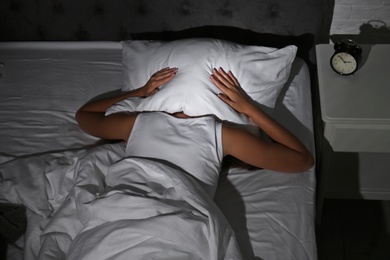 Photo of Young woman covering head with pillow in bed at home. Sleep disorder