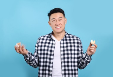 Photo of Asian man holding tasty fortune cookies with predictions on light blue background