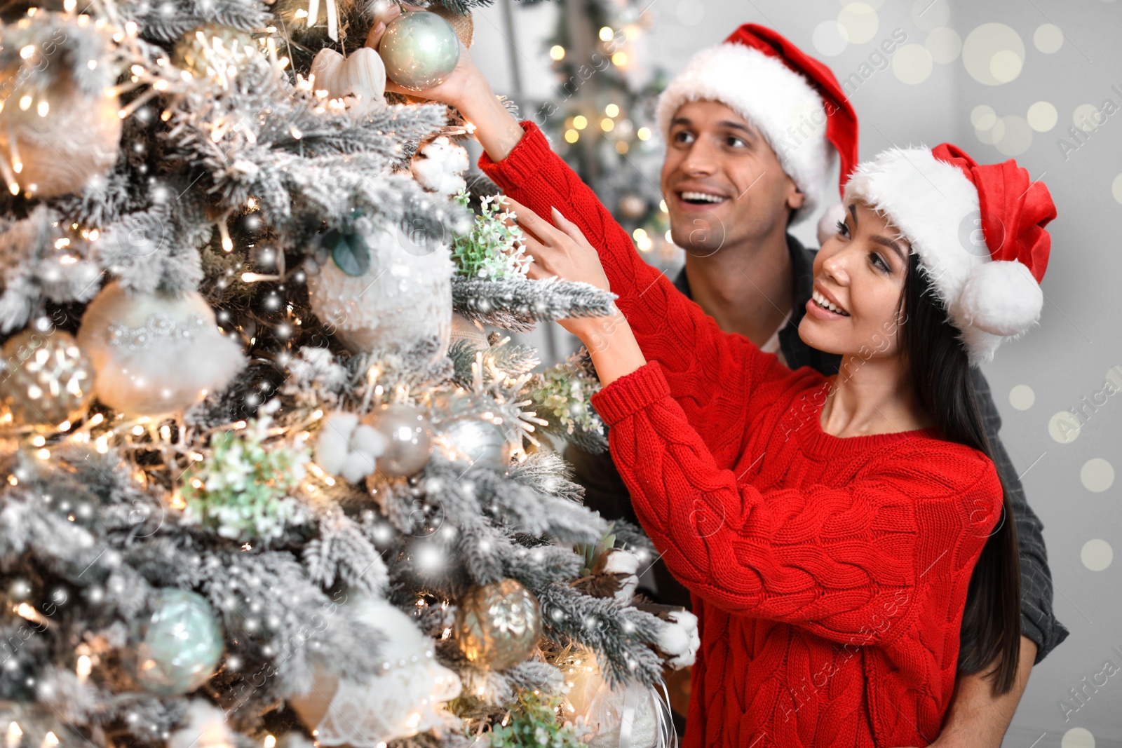 Image of Happy young couple in Santa hats decorating Christmas tree together at home