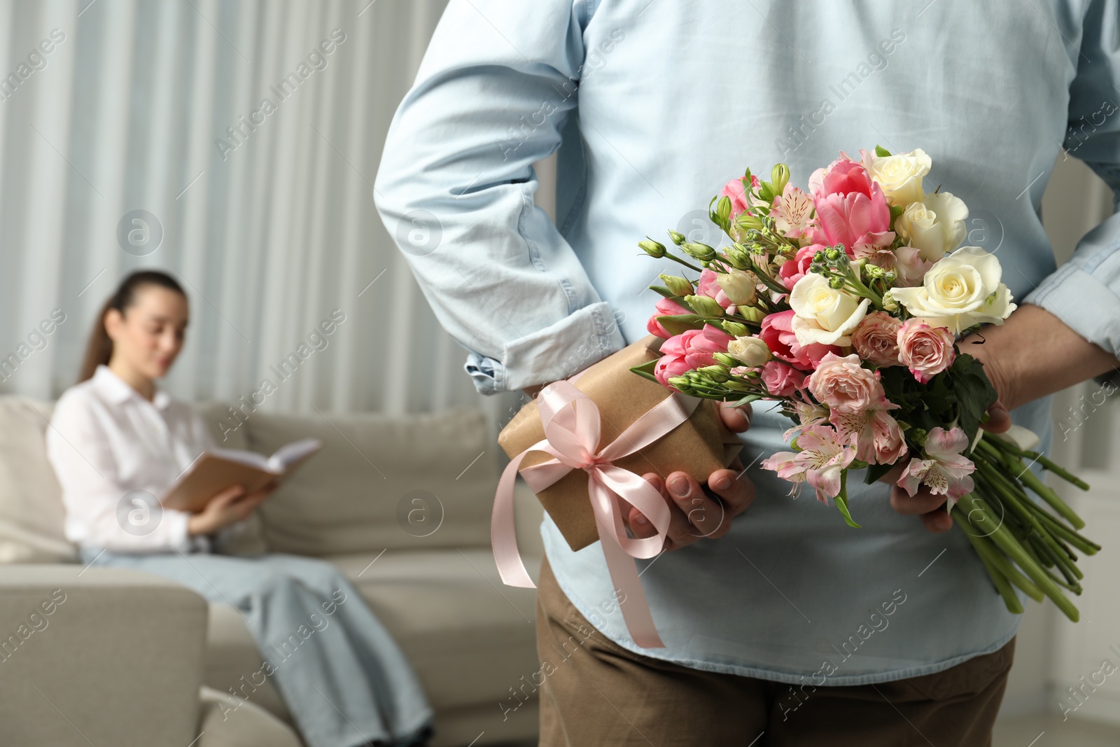 Photo of Man hiding bouquet of flowers and present for his beloved woman indoors, closeup