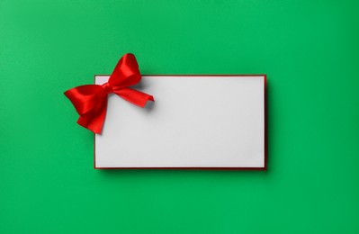 Photo of Blank gift card with red bow on green background, top view