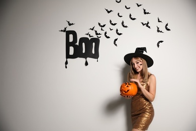 Woman in witch hat with jack o'lantern posing near white wall decorated for Halloween