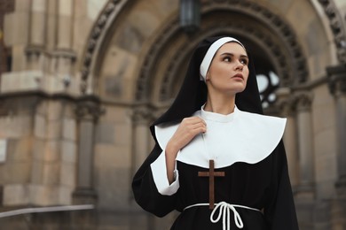 Young nun with Christian cross near building outdoors, space for text
