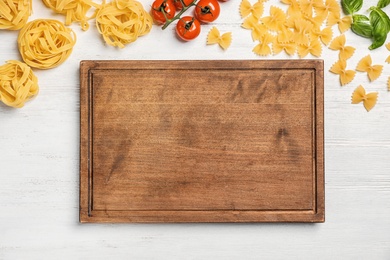 Wooden board and different raw pasta on table, top view