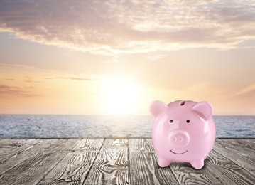 Image of Saving money for summer vacation. Piggy bank on wooden surface near sea at sunset, space for text