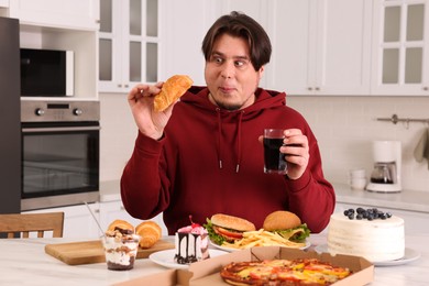 Photo of Overweight man with glass of cold drink and tasty croissant at table in kitchen