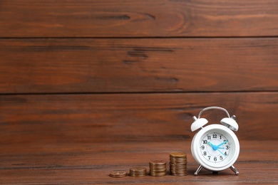 Photo of White alarm clock and stacked coins on table against wooden background, space for text. Money savings