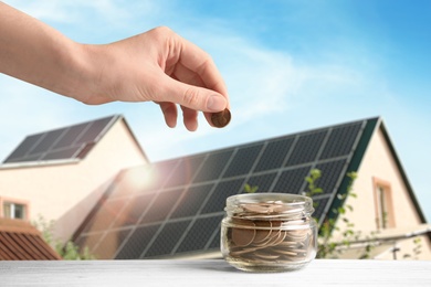 Image of Woman putting coin into jar against house with installed solar panels on roof, closeup. Economic benefits of renewable energy