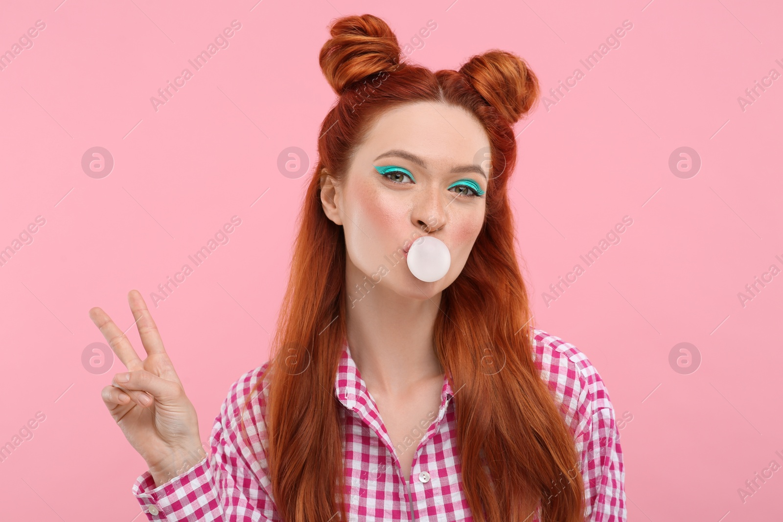Photo of Portrait of beautiful woman with bright makeup blowing bubble gum and showing peace gesture on pink background