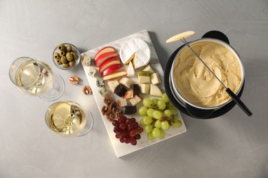 Photo of Fork with piece of apple, melted cheese in fondue pot, wine and snacks on grey table, flat lay