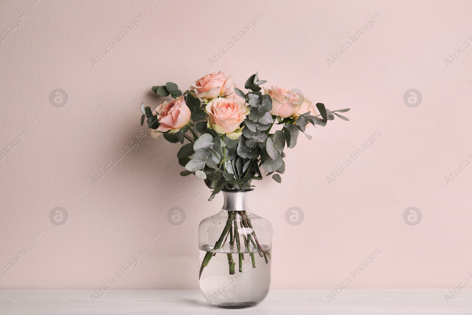 Photo of Vase with bouquet of beautiful roses on white wooden table near beige wall