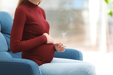 Photo of Young pregnant woman smoking cigarette at home, closeup. Harm to unborn baby