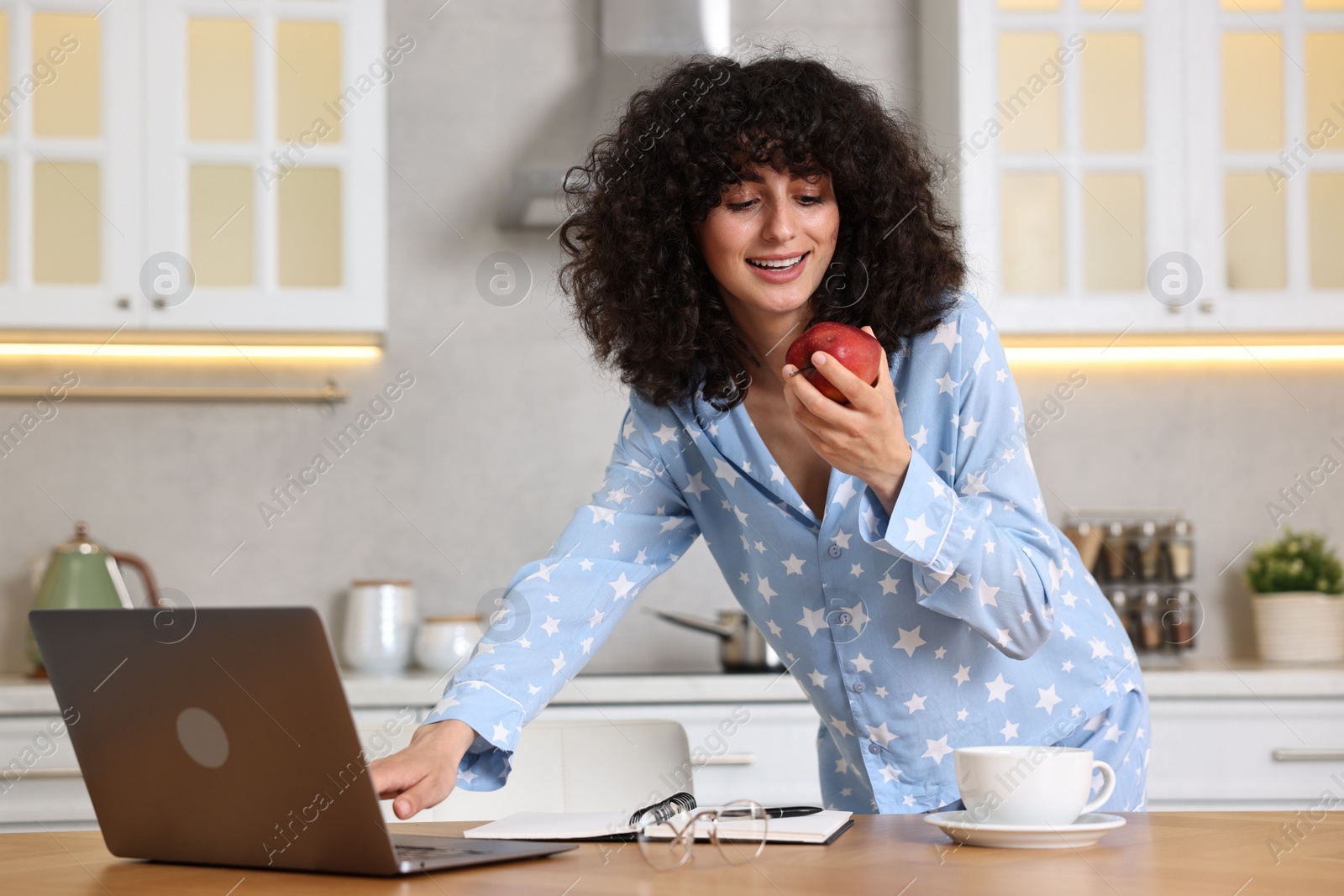 Photo of Beautiful young woman in stylish pyjama with fresh apple using laptop at wooden table in kitchen
