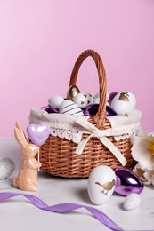 Wicker basket with festively decorated Easter eggs and bunny on white marble table against pink background