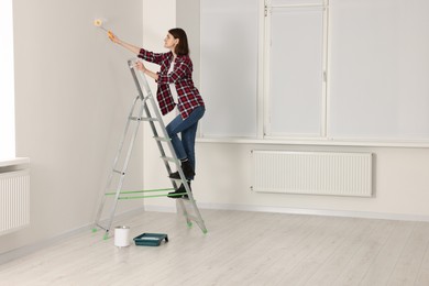 Photo of Young woman painting wall with roller on stepladder indoors. Room renovation