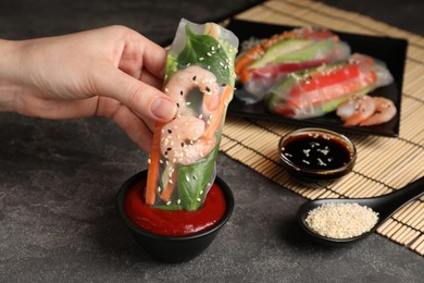 Woman dipping roll wrapped in rice paper into sauce at grey table, closeup