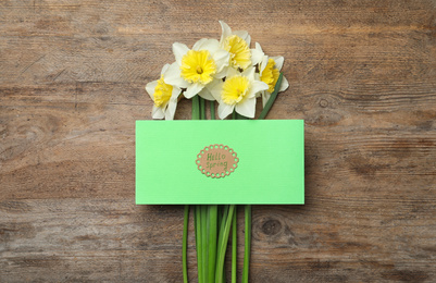 Green card with words HELLO SPRING and narcissus on wooden table, flat lay