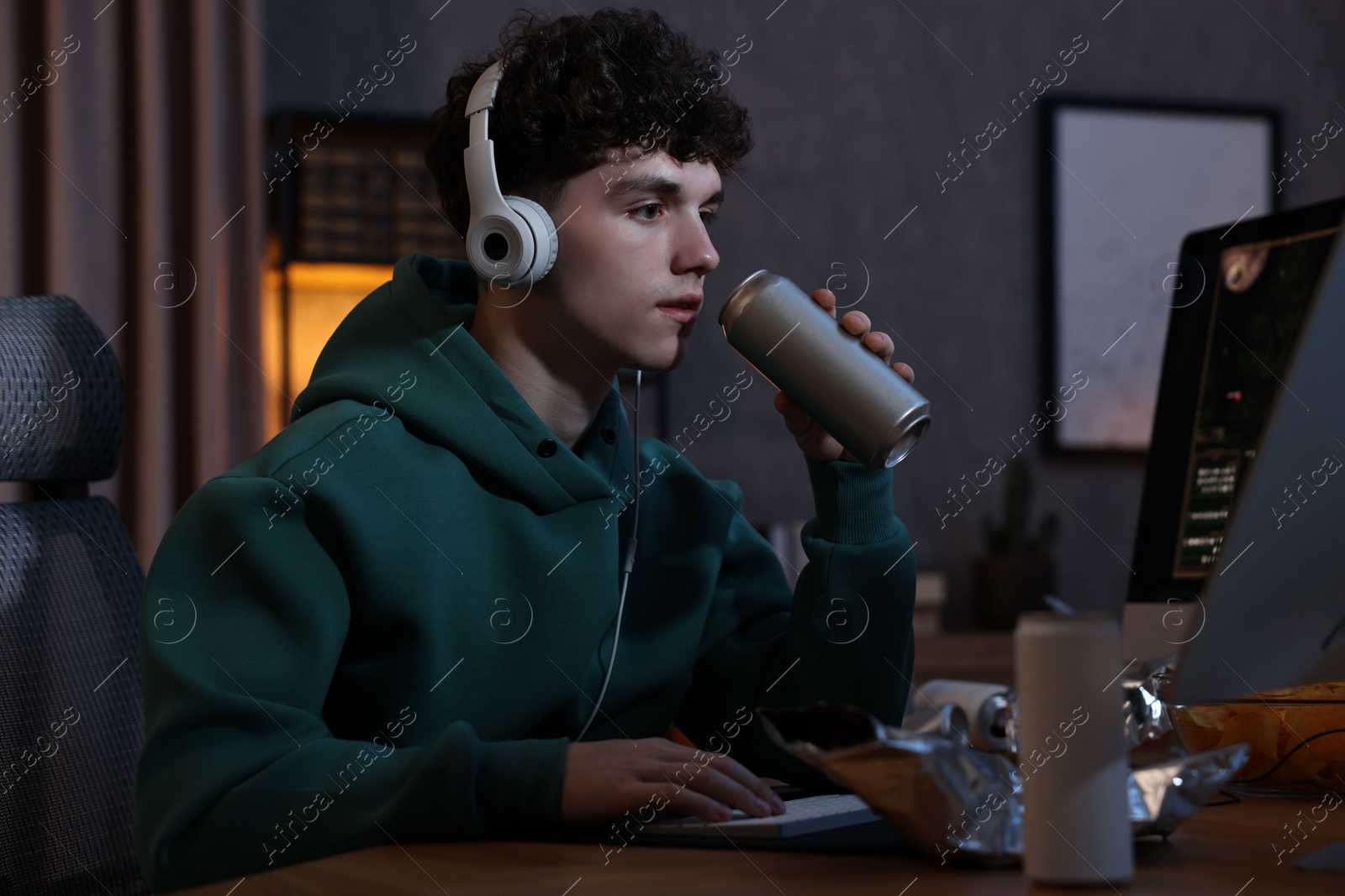 Photo of Young man with energy drink and headphones playing video game at wooden desk indoors