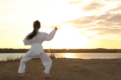 Photo of Cute little girl in kimono practicing karate near river at sunset, back view