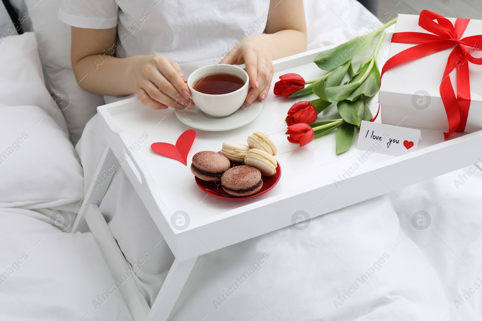 Photo of Tasty breakfast served in bed. Woman with tea, macarons, gift box, flowers and I Love You card at home, closeup