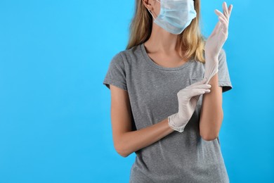 Photo of Young woman in protective mask putting on medical gloves against light blue background, closeup. Space for text
