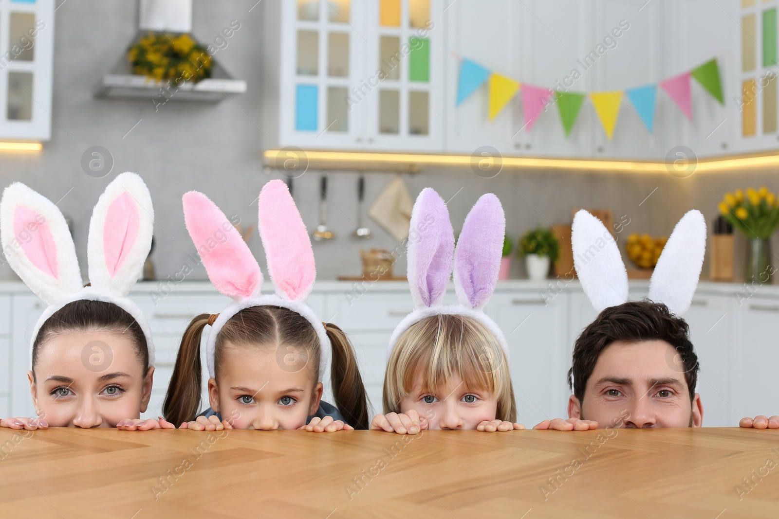Photo of Happy family wearing bunny ears headbands and peeking over table in kitchen. Easter celebration