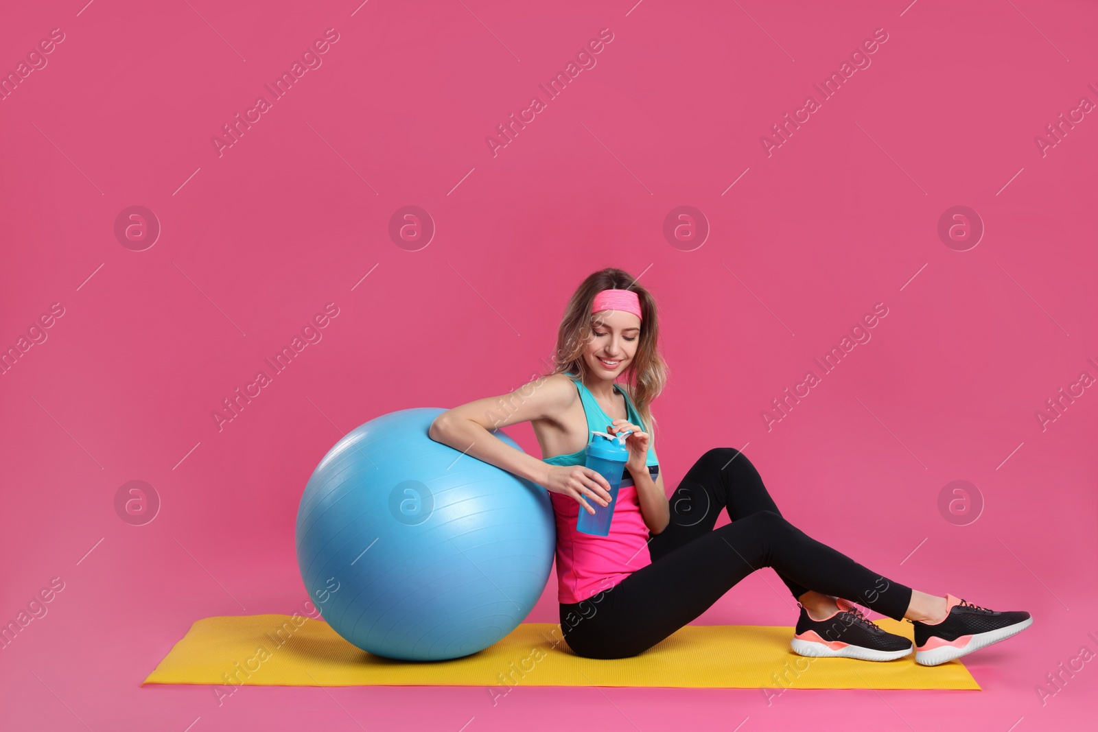 Photo of Beautiful woman sitting on yoga mat with fitness ball against pink background